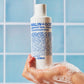 MALIN+GOETZ grapefruit face cleanser held by a hand with soap suds against a tiled wall background