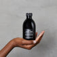 Hand holding a black bottle of OI Body Wash against a gray background