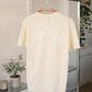 Beige GOAT x Harvey Nichol Tee hanging on a clothes rack with home objects on a shelf in the background