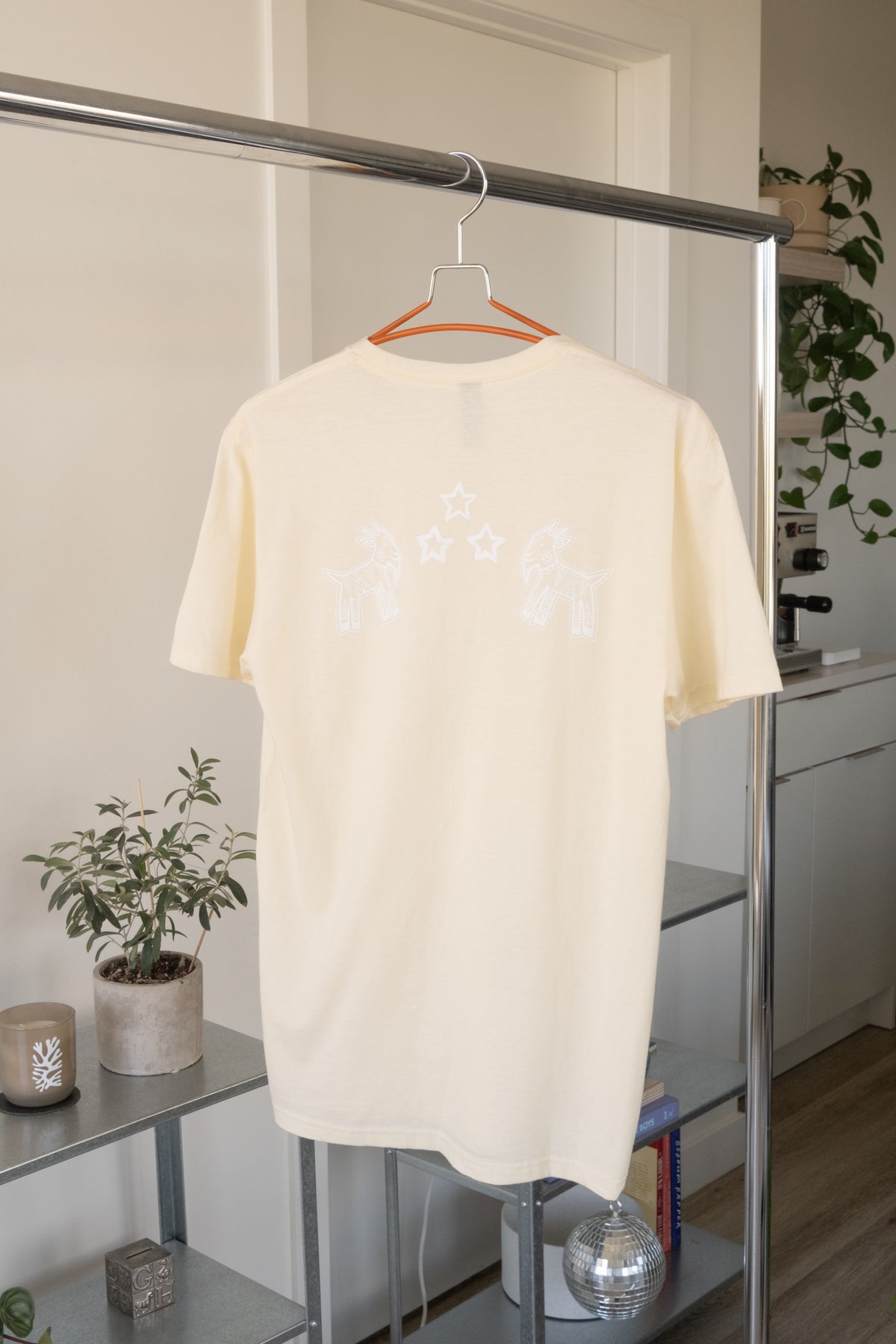 Beige GOAT x Harvey Nichol Tee hanging on a clothes rack with home objects on a shelf in the background