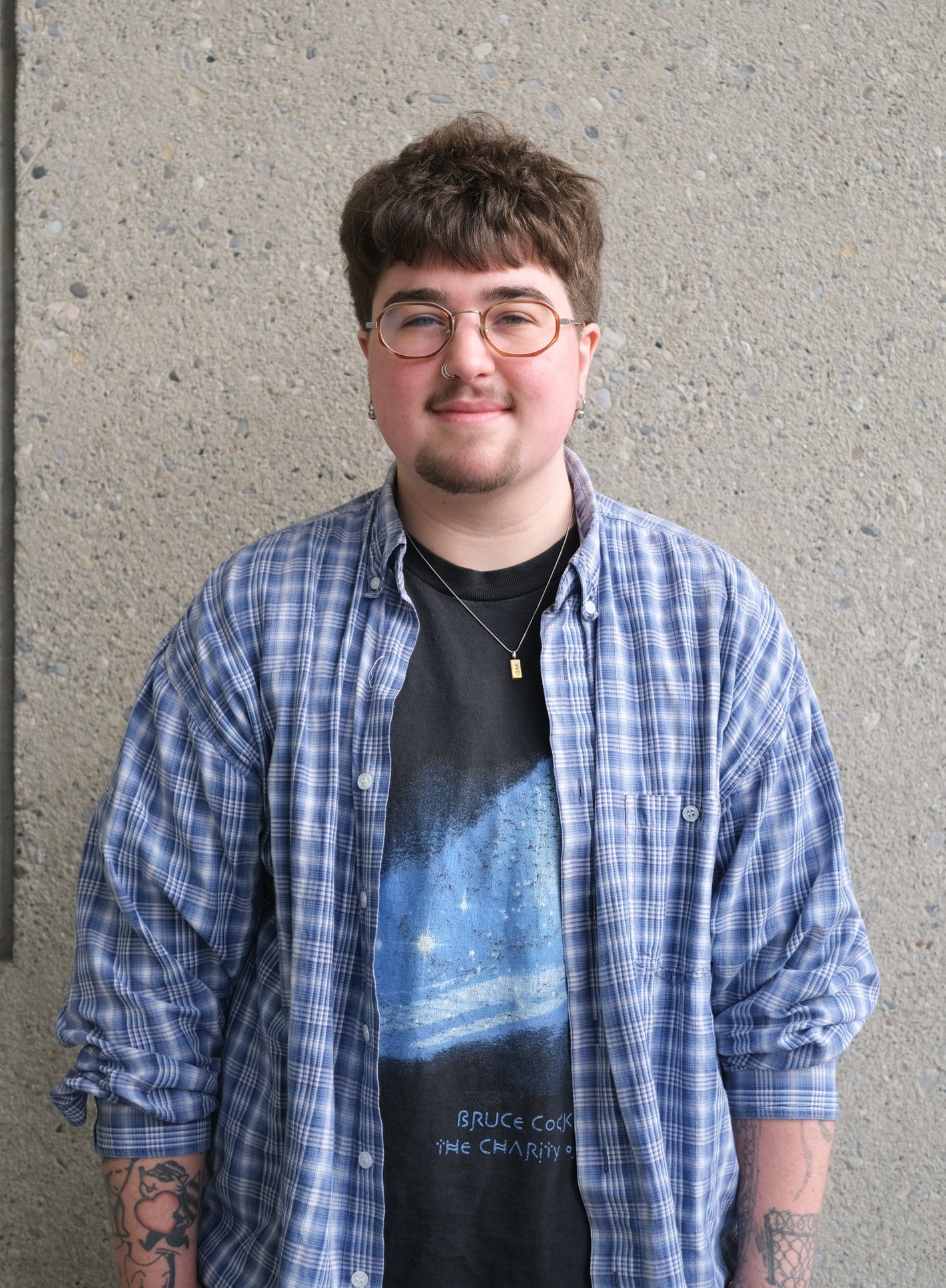 Person wearing a blue plaid shirt over a black graphic t-shirt against a gray wall.
