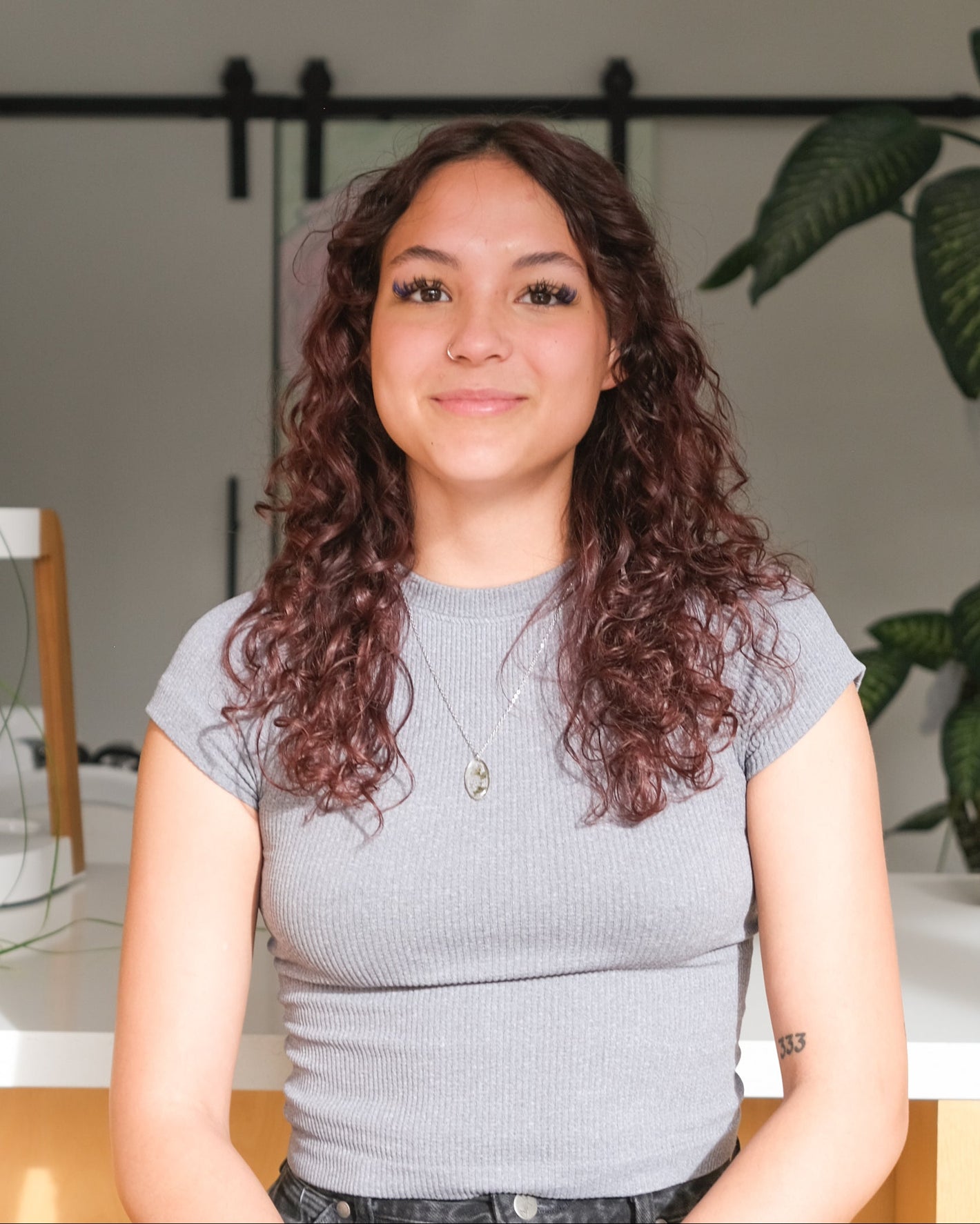 Woman with curly hair wearing a gray shirt in a modern indoor setting with a big houseplant in the background.