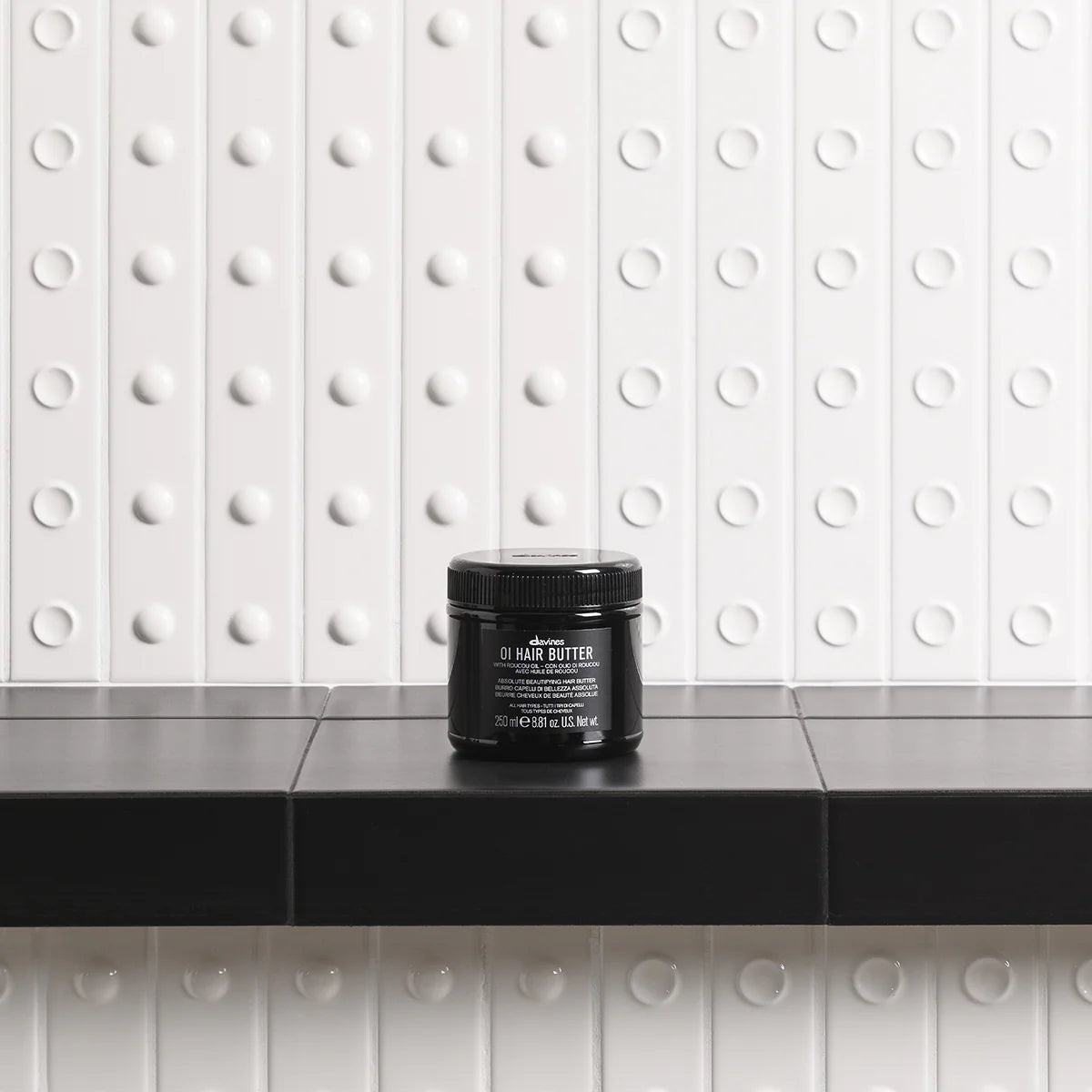 Black jar of hair butter on a black countertop with a white tiled wall background