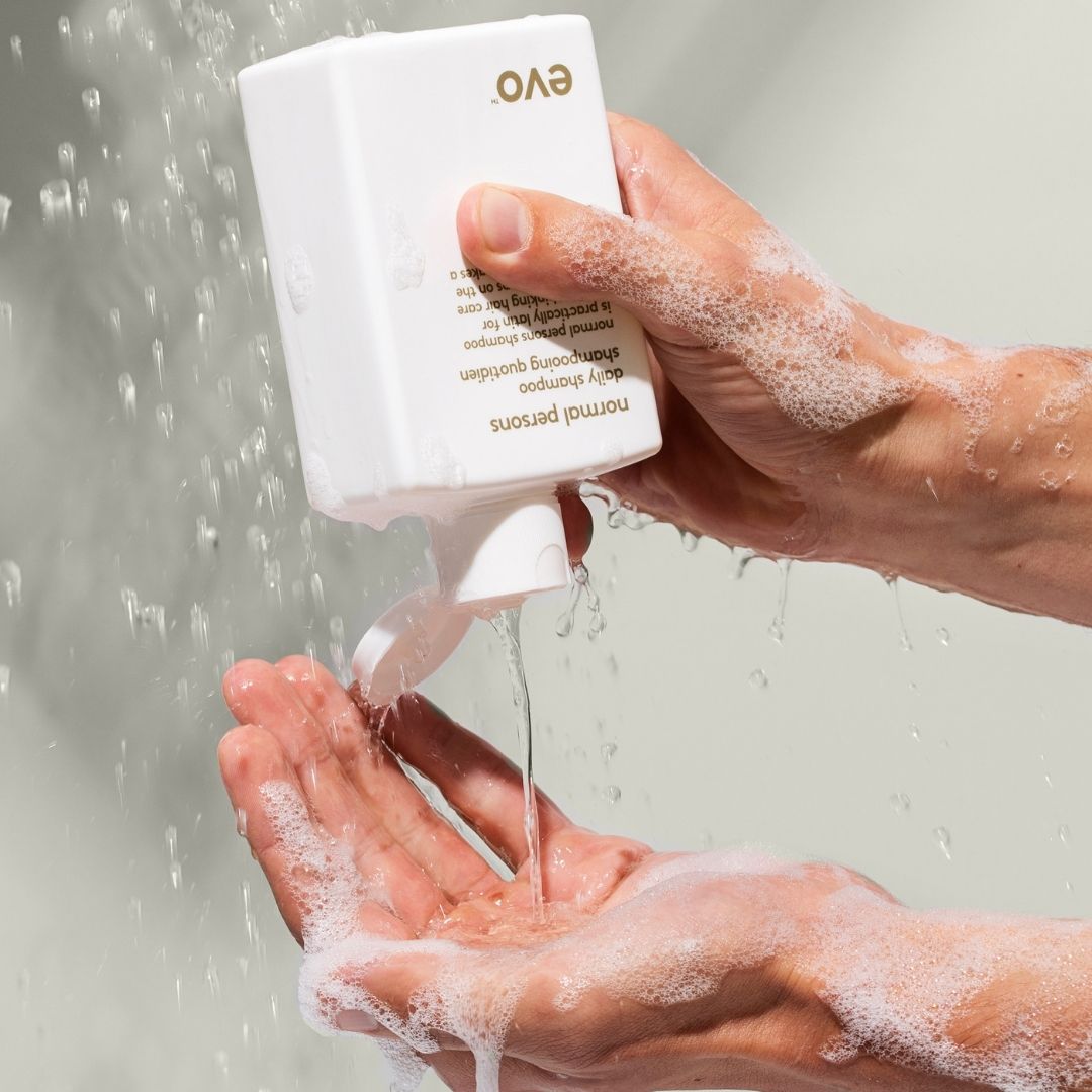 Hand holding a white bottle labeled 'Evo' under running water, with soap suds on hands and bottle.