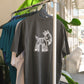 Dark grey GOAT x Harvey Nichol t-shirt hanging on a clothes rack with a green plant in the back