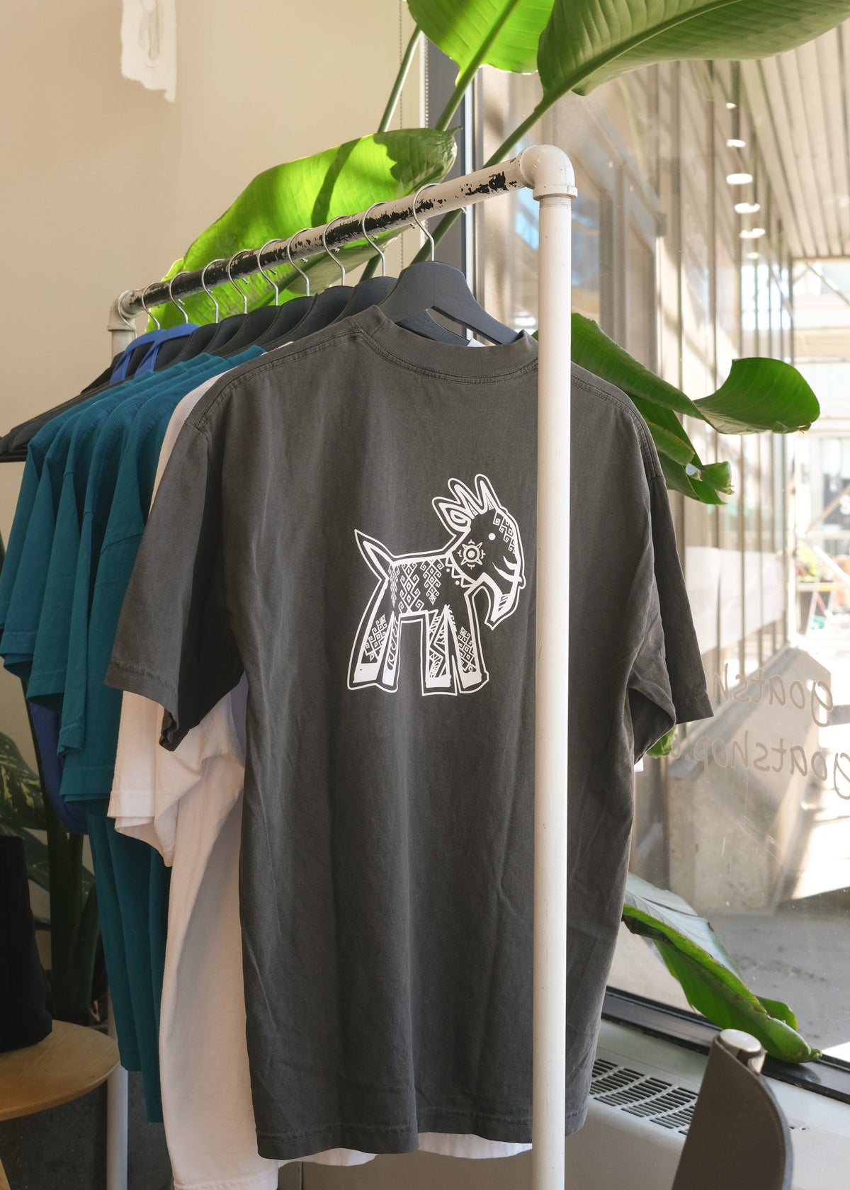 Dark grey GOAT x Harvey Nichol t-shirt hanging on a clothes rack with a green plant in the back