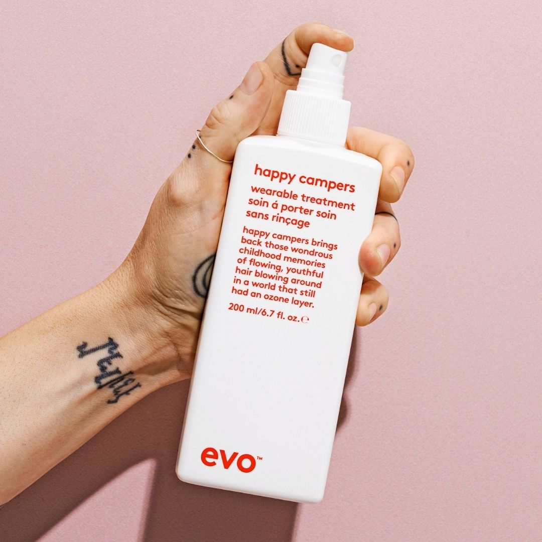 Hand holding a bottle of 'happy campers' treatment by evo against a pink background