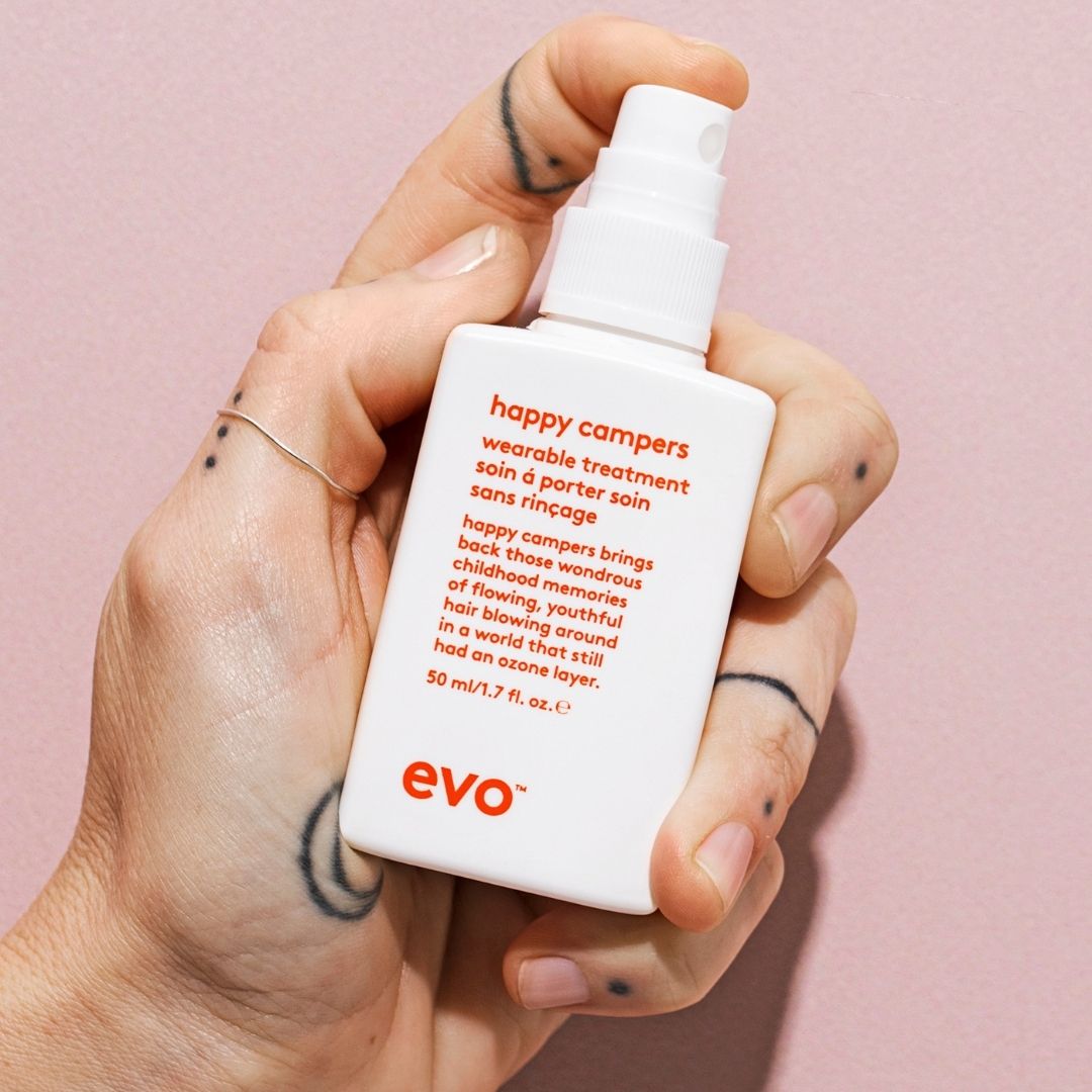 Hand holding a white bottle labeled 'happy campers' by evo against a pink background