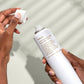 Person holding a white spray bottle with text, against a neutral background