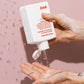 Hand holding a white bottle labeled 'EVO' with shampoo being dispensed onto a palm against a pink background
