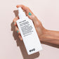 Hand holding a white bottle of 'root canal' volumizing spray by evo against a light pink background