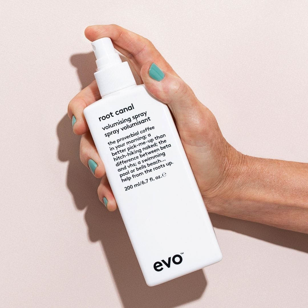 Hand holding a white bottle of 'root canal' volumizing spray by evo against a light pink background