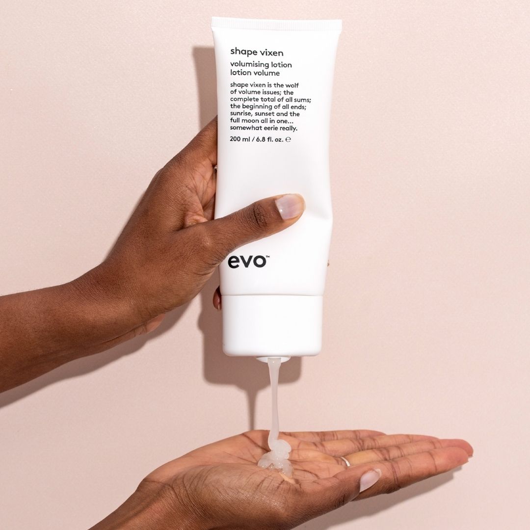 Hand holding a white bottle of evo 'shape vixen' volumizing lotion against a beige background