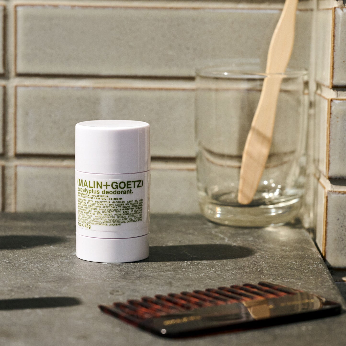 Malin+Goetz deodorant stick on a bathroom counter with a glass and comb.