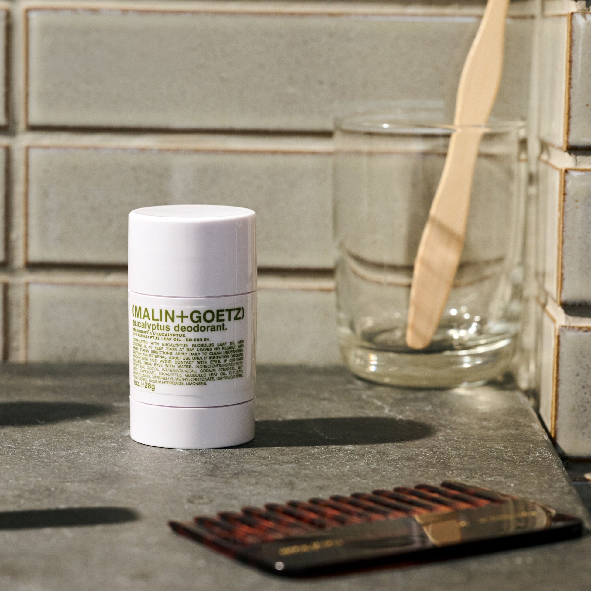 Malin+Goetz deodorant stick on a bathroom counter with a glass and comb.