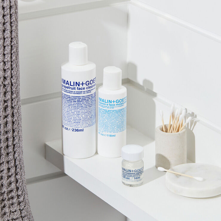 MALIN+GOETZ skincare products on a bathroom shelf with a towel and toothbrush holder.